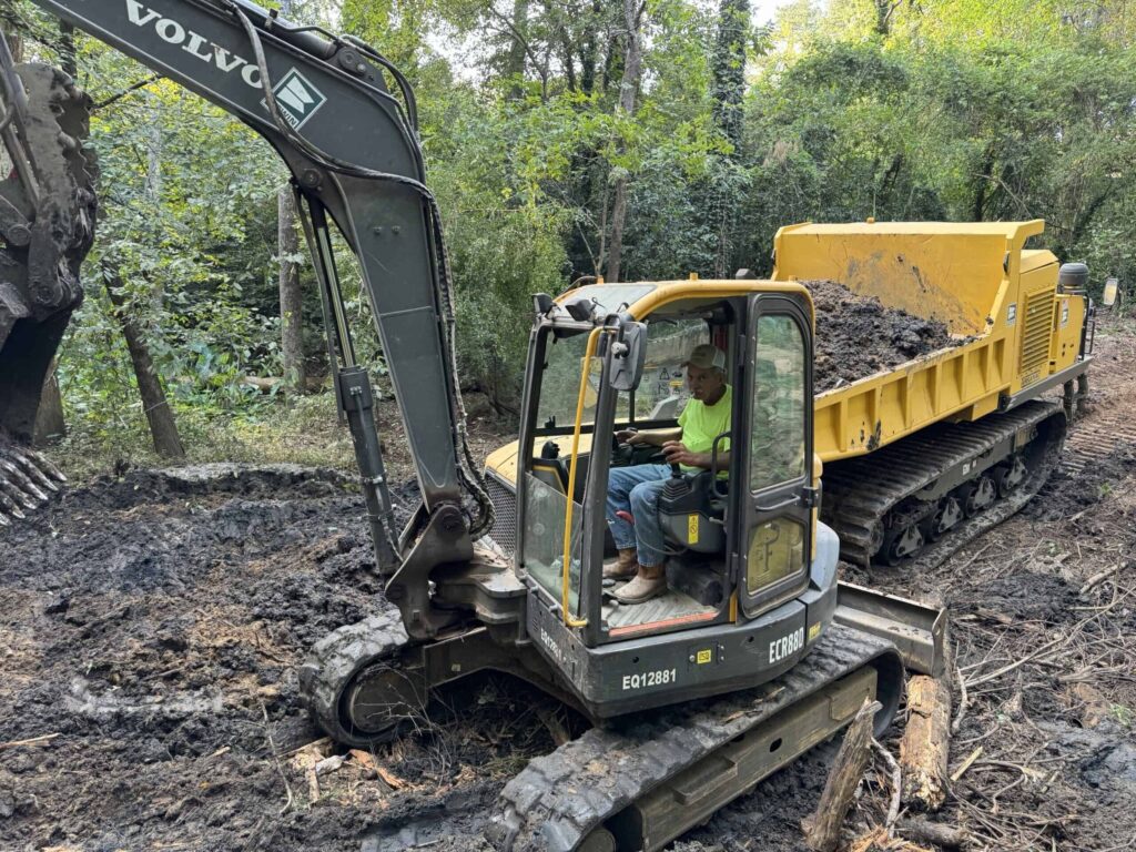 Excavator operator working on a muddy terrain in a forested area.
