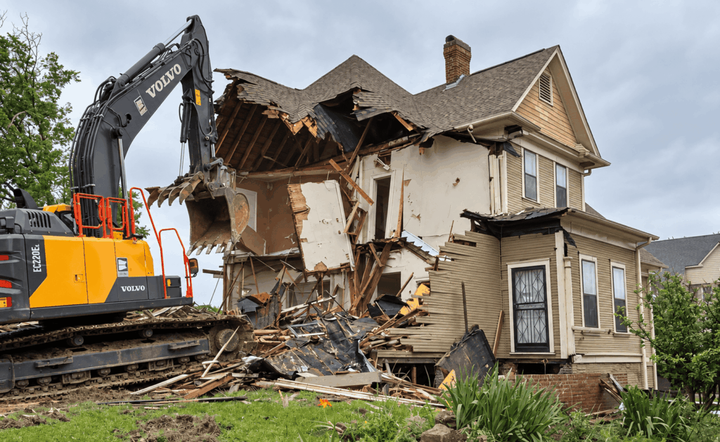 Excavator demolishing a house, debris scattered around.